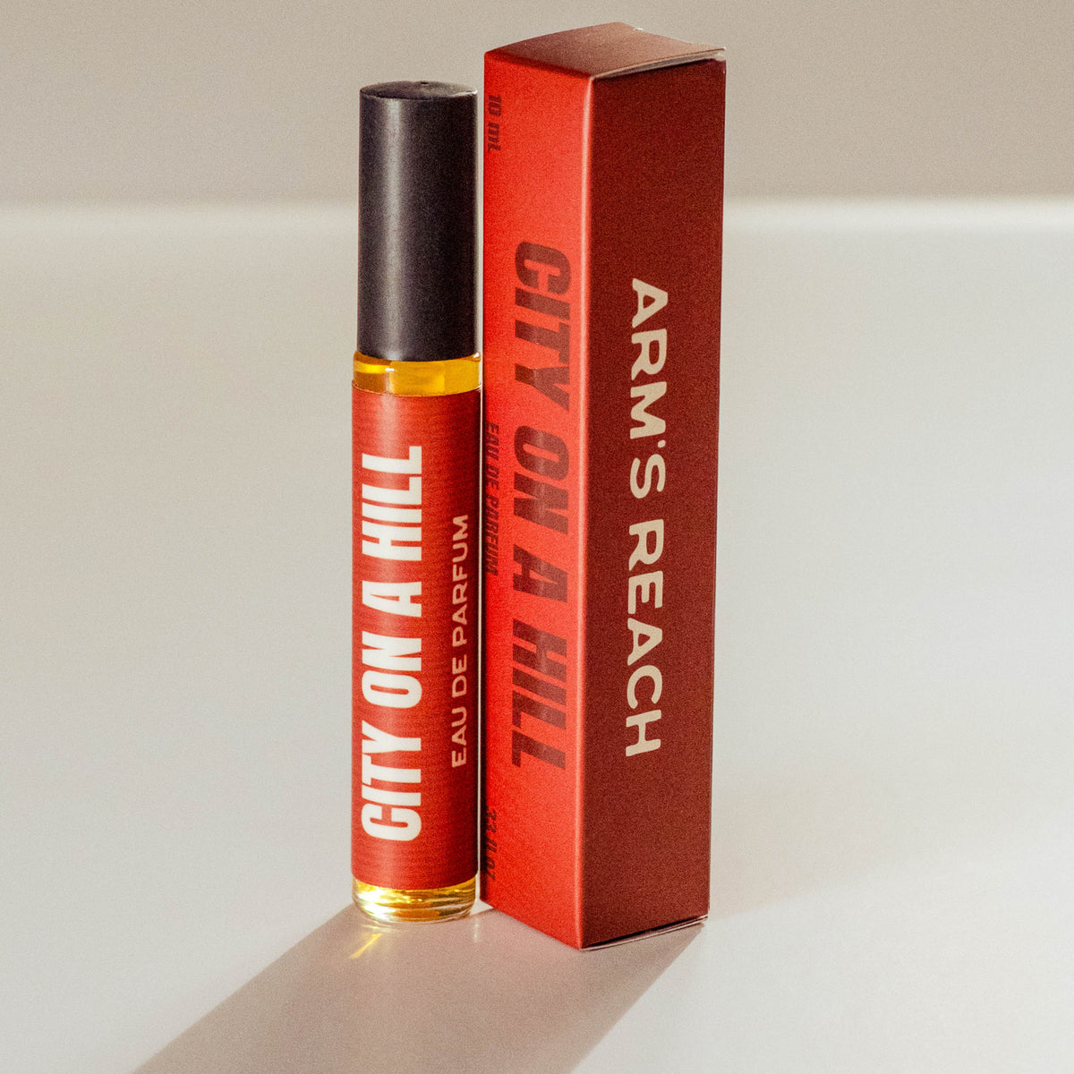 A small perfume bottle with a black cap stands next to its red box. The bottle and box are both labeled 'CITY ON A HILL' and 'ARMS REACH', with 'EAU DE PARFUM' printed below the title.