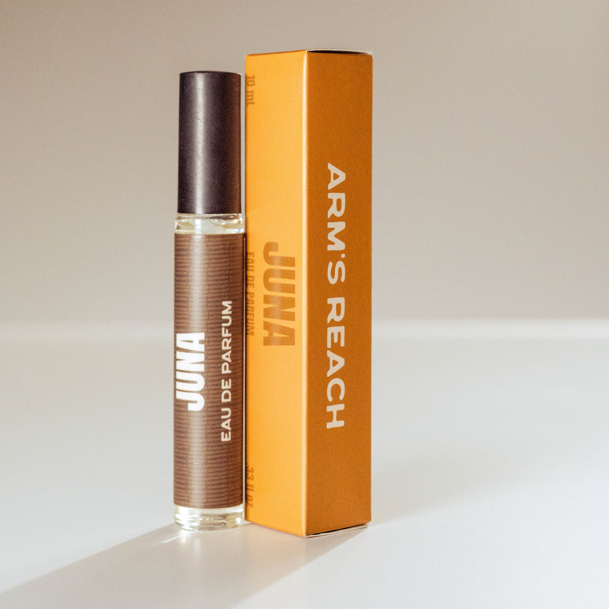 A bottle of Juna Eau de Parfum with a dark brown cap and a brown textured label, standing next to its orange cardboard packaging box on a light gray background.
