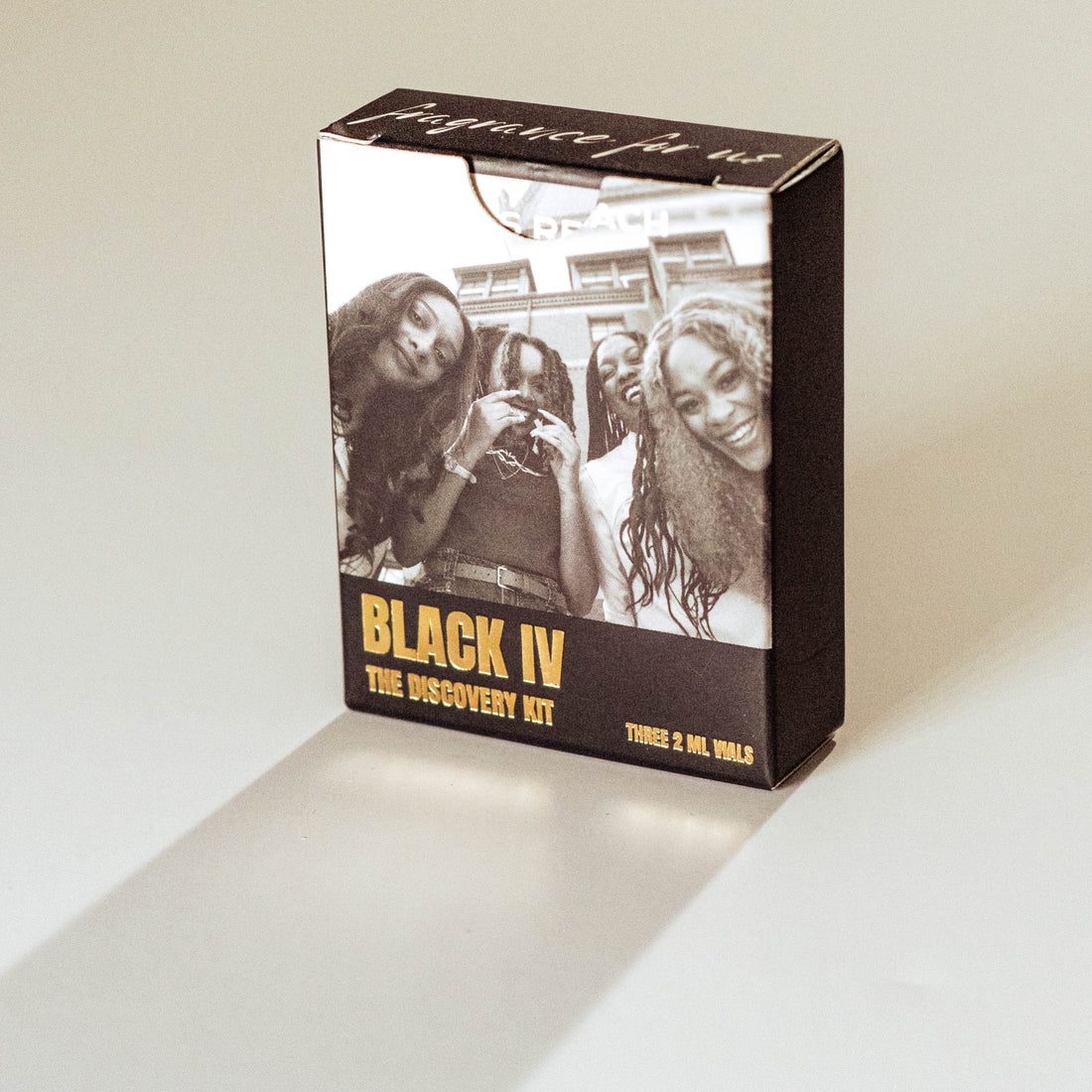 A rectangular product box standing upright on a light beige surface, featuring a black-and-white photo of three women on the front with the text 'BLACK IV THE DISCOVERY KIT' in yellow and white. The box is photographed in a clean studio setting with a soft shadow cast to the lower left.