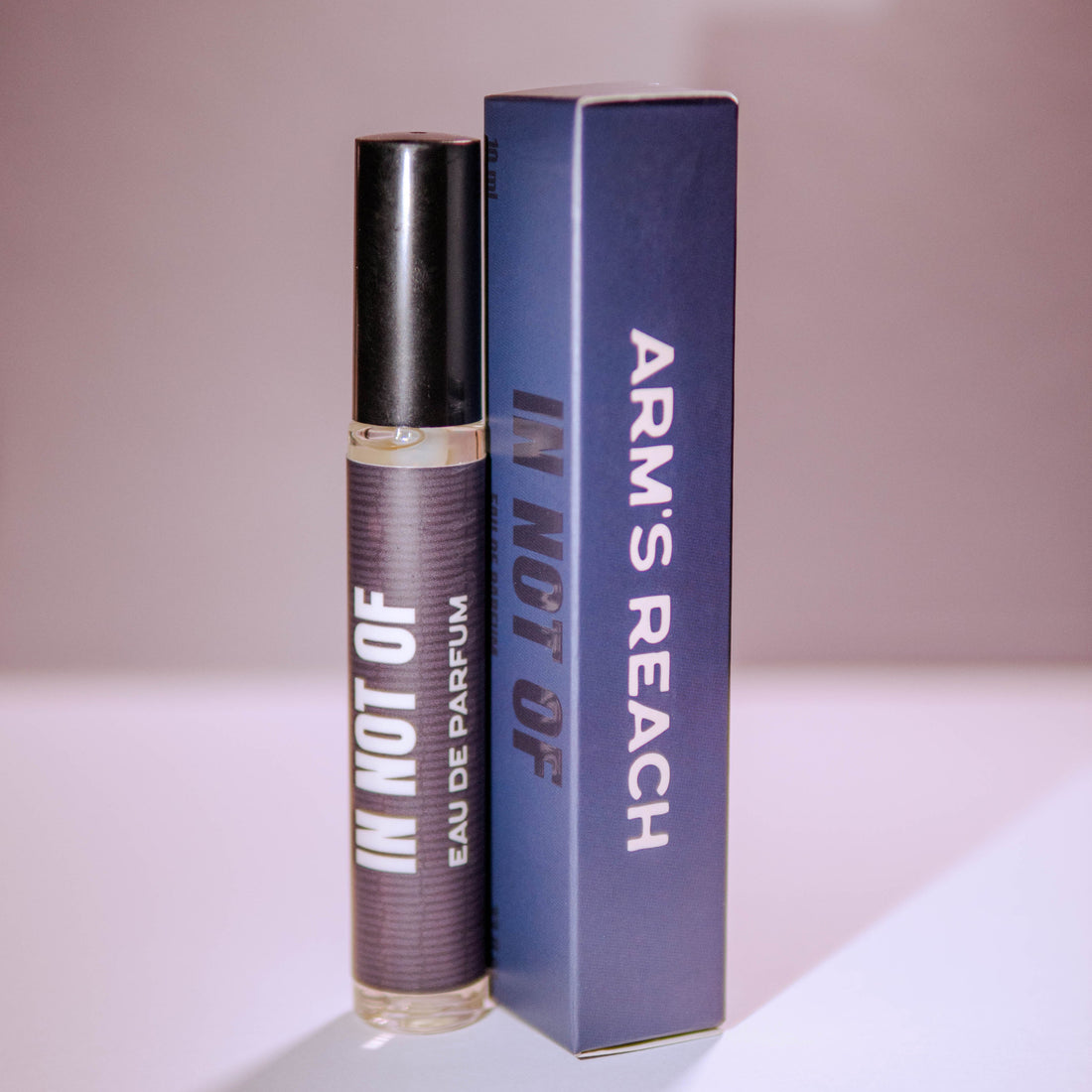 A perfume bottle with a black cap stands next to its blue box. The bottle label reads 'IN NOT OF EAU DE PARFUM' and the box displays 'ARM'S REACH' and 'IN NOT OF' in white text.