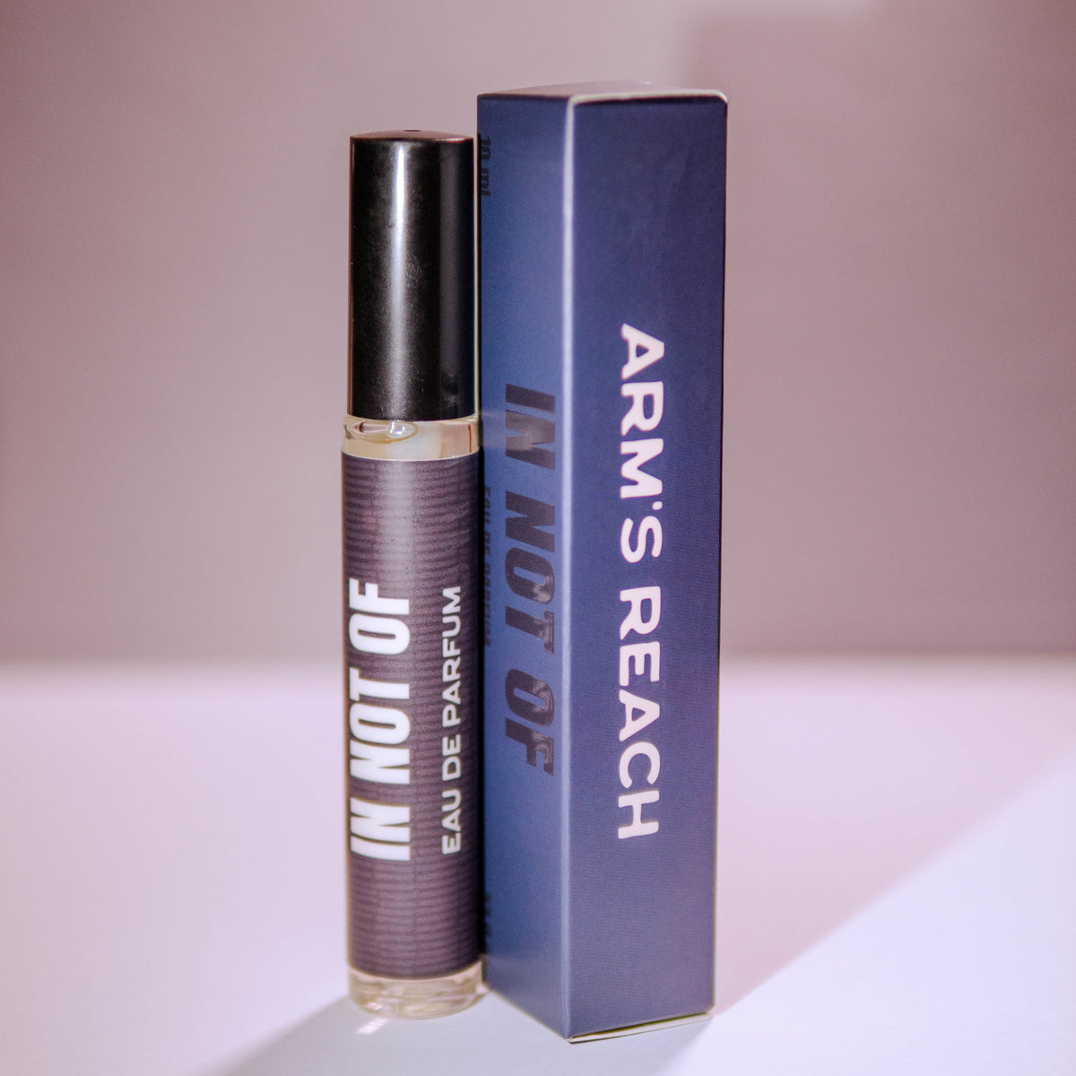 A perfume bottle with a black cap stands next to its blue box. The bottle label reads 'IN NOT OF EAU DE PARFUM' and the box displays 'ARM'S REACH' and 'IN NOT OF' in white text.