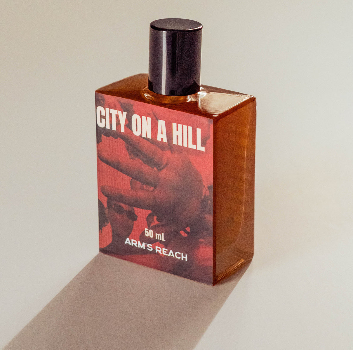 A 50ml bottle of Arm's Reach 'City on a Hill' perfume, featuring a translucent amber-brown glass body, a red graphic label, and a black cap, set against a plain light beige background with a soft shadow.