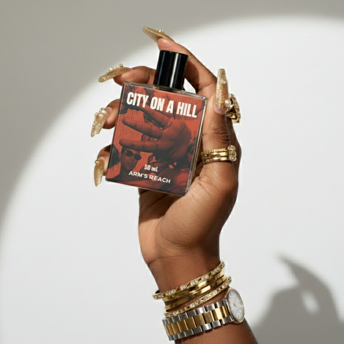 Hand holding a 'City on a Hill' perfume bottle with a red label against a white background