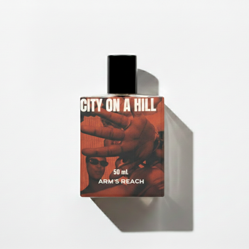 Perfume bottle with 'City on a Hill' packaging featuring a hand and sunglasses on a light gray background